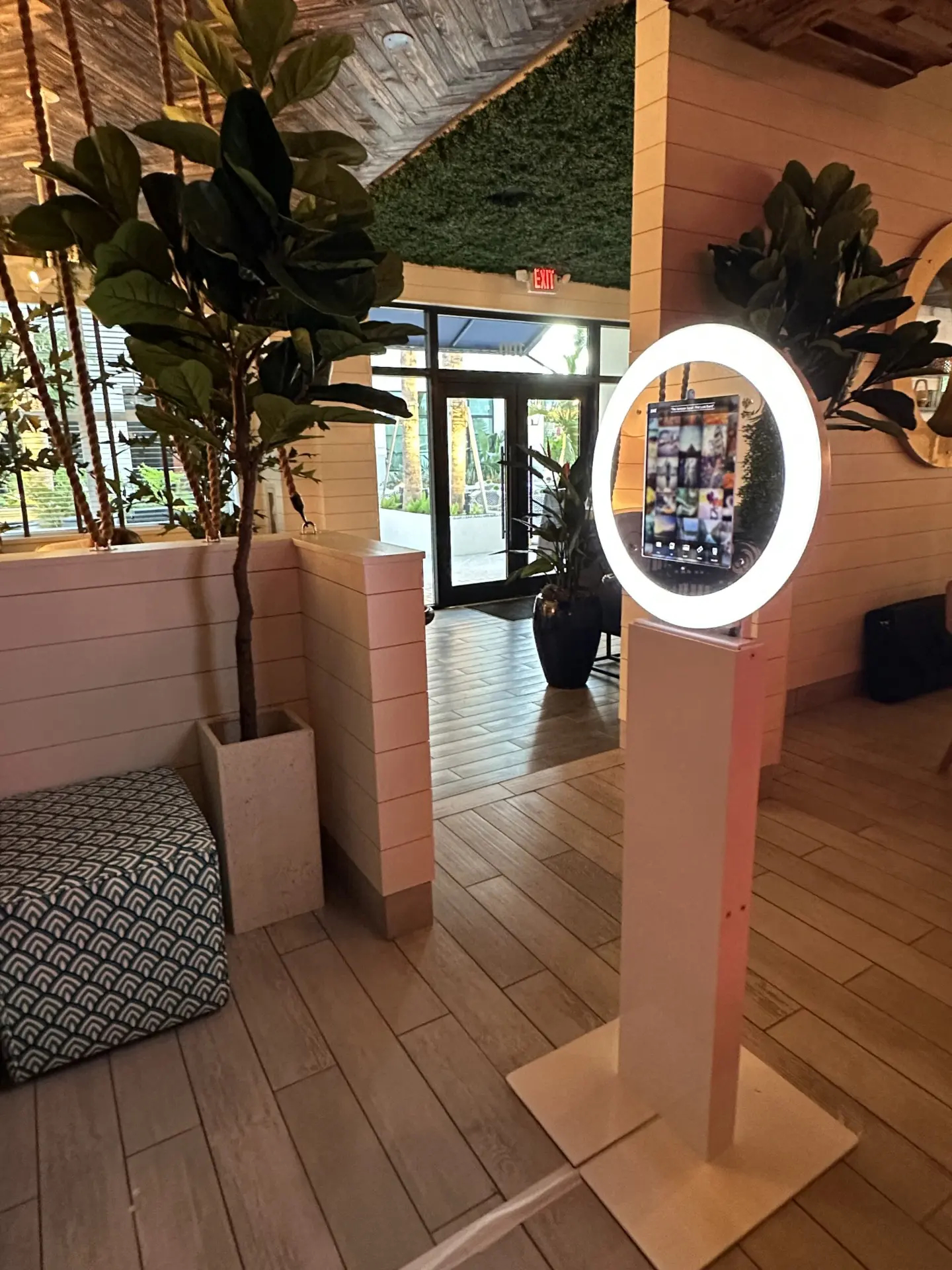 White digital photo booth with a glowing ring light standing in a warm venue entrance beside lush plants